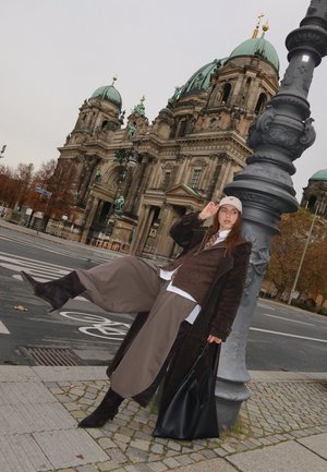 Model wearing a brown knitted vest over a white shirt, gray wide-leg pants, and black knee-high boots, holding a black bag, in front of a historic building.