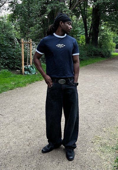 Camiseta azul marino con detalles en blanco y texto "NYC", combinada con jeans negros holgados y zapatos negros. Entorno exterior con vegetación.