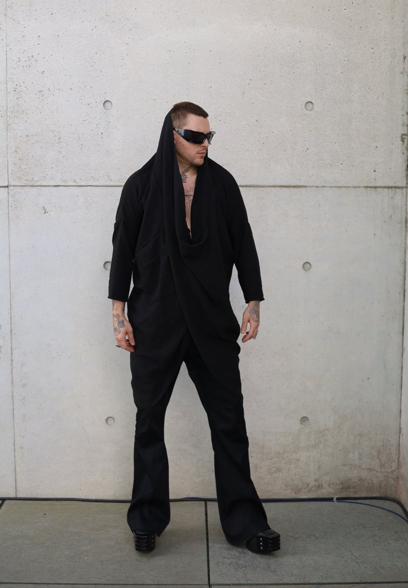 Μαύρη υφασμάτινη εμφάνιση με σχέδιο κατρακύλας, που διαθέτει λαιμόκοψη καβάλου και χαλαρή εφαρμογή, συμπληρωμένη με χοντρές μαύρες τσάντες και γυαλιά ηλίου.