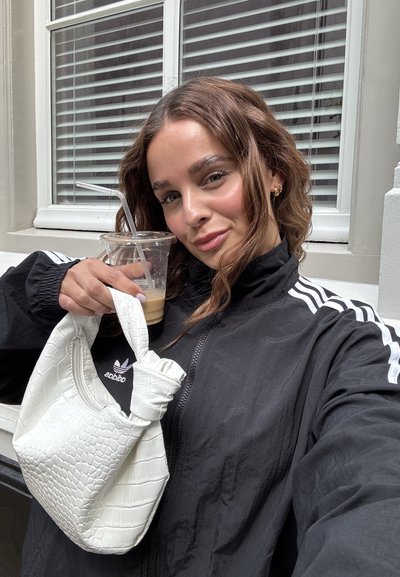 Bolso de mano de imitación de cocodrilo blanco con un asa anudada, sostenido junto a un vaso transparente con una pajita, contra un fondo de listones de ventana.