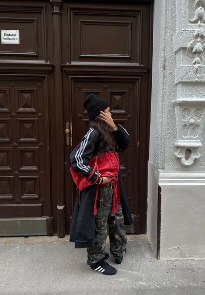 Gorro negro, abrigo largo negro con rayas blancas, pantalones de camuflaje con patrón, zapatos negros, llevando una bolsa roja de cuerda, de pie junto a puertas marrones.