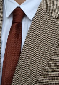 Blazer marrone con motivo pied-de-poule e risvolti testurizzati, abbinato a una camicia bianca e una cravatta tinta unita color ruggine.