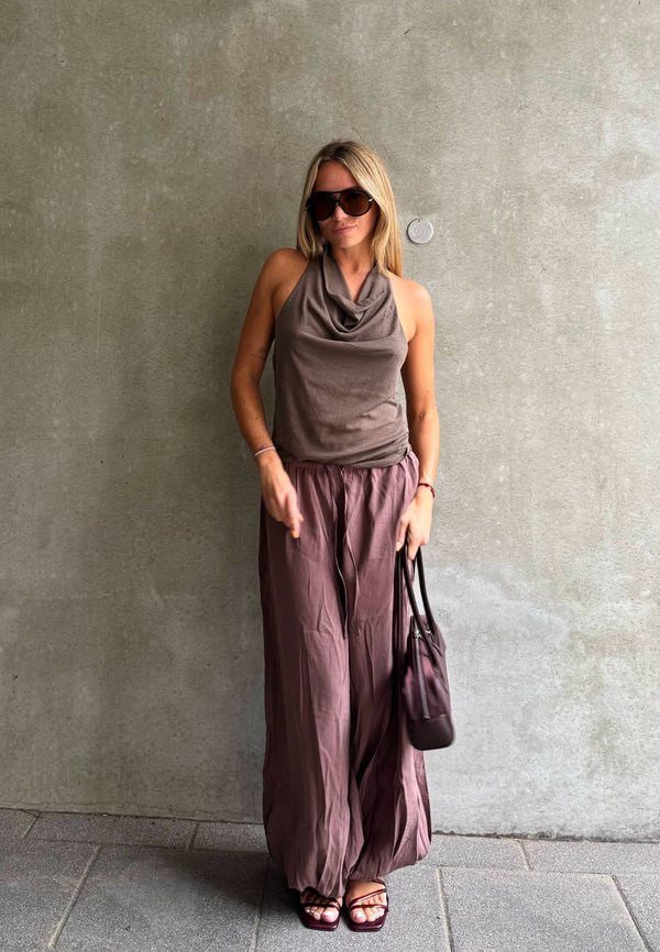 Mujer con gafas de sol, top sin mangas, pantalones anchos y sandalias, sosteniendo un bolso, de pie frente a una pared de hormigón lisa.