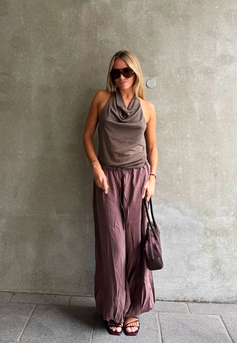 Femme portant des lunettes de soleil, un haut sans manches, un pantalon large et des sandales, tenant un sac à main, debout contre un mur en béton uni.