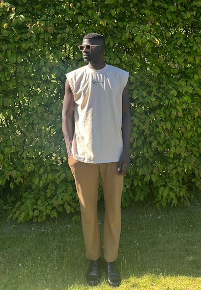 Camisa sin mangas de gran tamaño en color beige con logo negro, pantalones de sastre en tono marrón y botas negras, todo sobre un fondo de hojas verdes.