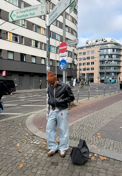 Chaqueta bomber de piel negra, jeans de mezclilla azul claro de corte oversized con un parche rojo, zapatos marrones y una bolsa oscura en una calle de adoquines.