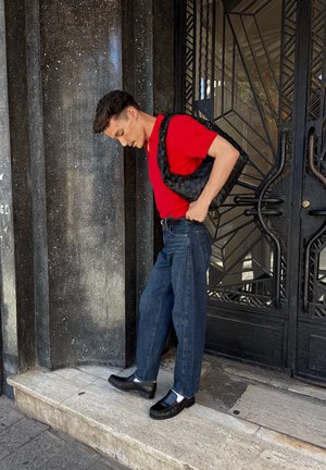 Joven con camisa roja y vaqueros de pie sobre un escalón de mármol, ajustándose los pantalones con un bolso negro tejido al hombro junto a una puerta de metal negra.