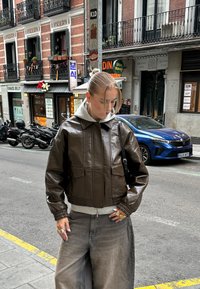 Brun læder cropped jakke med krave, to frontlommer, elastiske manchetter og en grå hoodie lagdelt under. Bred grå bukser.