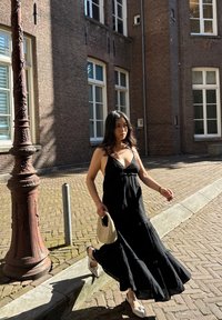 Schwarzes Maxikleid mit gestuften Design und Spaghettiträgern. Auffällige gewebte beige Handtasche und spitze Schuhe mit Verzierungen. Ziegelhintergrund.