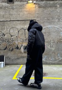 Fekete pufi dzseki szőrmebéléssel ellátott kapucnival, laza fekete nadrággal és fekete kesztyűvel párosítva. Szürke fal graffitivel és sárga jelekkel.