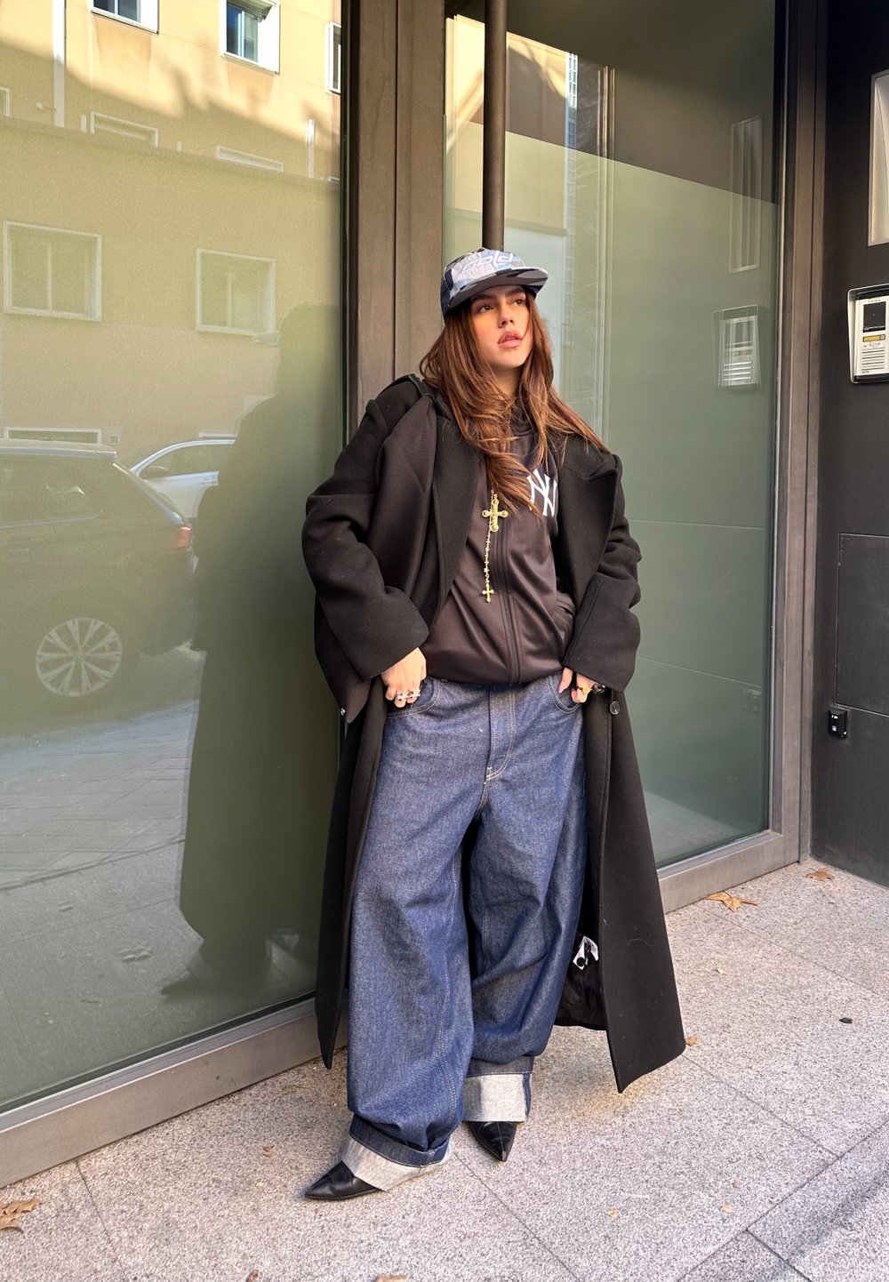 Μαύρο παλτό, γκρίζο καπέλο, μαύρη μπλούζα με λογότυπο, χρυσή αλυσίδα με σταυρούς, φαρδιά μπλε τζιν με μανσέτες, μαύρα μυτερά παπούτσια, έρχεται κοντά σε γυάλινο τοίχο.
