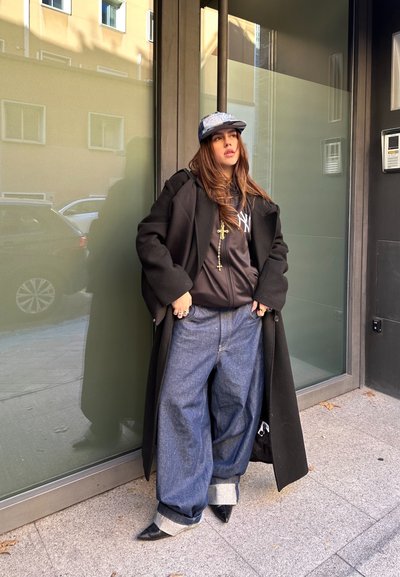 Abrigo negro, gorra gris, sudadera negra con logo, cadena dorada con cruces, jeans azules holgados con dobladillos, zapatos negros de punta, de pie junto a una pared de cristal.