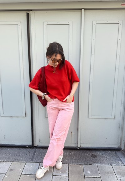 Mujer con camisa roja, pantalones rosas, zapatillas blancas y gafas de sol, de pie en la acera frente a una puerta gris con las manos en los bolsillos.