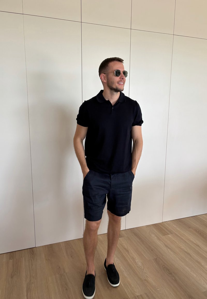 Homme portant un polo noir, un short foncé et des lunettes de soleil, debout les mains dans les poches sur un sol en bois près d'un mur blanc à panneaux.