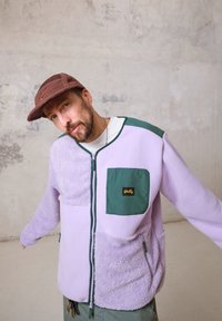 Casaco de fleece em lavanda com detalhes verdes, apresentando bolsos com fecho éclair, painéis texturizados e um logo em destaque na frente.