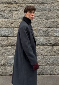 Lange donkergrijze jas met een hoge kraag, gedragen met bordeauxrode handschoenen. Op de achtergrond een getextureerde stenen muur. Horizontale weergave van de jas.