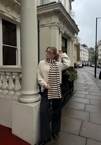 Maglione beige e pantaloni neri a vita alta, abbinati a una sciarpa a righe nera e beige. Lo sfondo presenta un edificio storico e una strada.