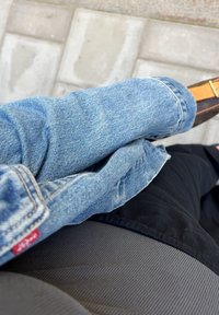 Μπλε τζιν παντελόνι με ξεθωριασμένη υφή, με τυλιγμένα μπατζάκια και κόκκινο ταμπελάκι, τοποθετημένο πάνω από μαύρο ύφασμα διαφορετικής υφής και χρώματος.