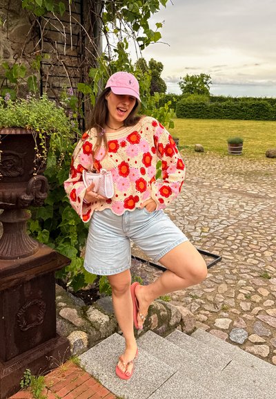 Joven mujer que lleva un suéter rosa con estampado floral, pantalones cortos de mezclilla, gorra rosa y chanclas, posando al aire libre cerca de plantas y un camino de piedra.