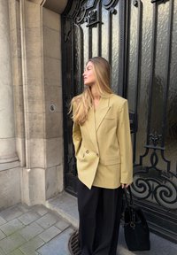 Blazer croisé vert olive avec des revers marqués, doté de poches sur la poitrine et sur les côtés, associé à un pantalon noir à jambes larges et à un sac à main noir.