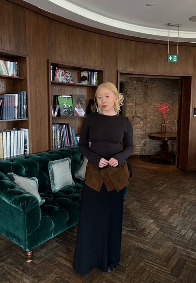 Top de manga larga negro, detalles de envoltura de ante marrón, falda maxi negra. Sofá de terciopelo verde oscuro con almohadas decorativas, acentos de interior de madera.