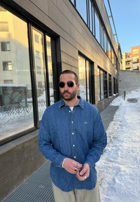 Denim hosszú ujjú ing, gombos dizájnnal, kék színben, logóval a mellkason; ezüst gyűrűkkel és kerek napszemüveggel kiegészítve. Havas háttér.