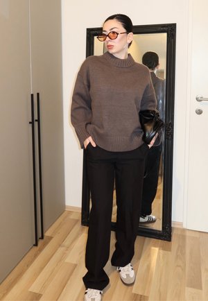 Mujer con suéter marrón, pantalones negros, zapatillas blancas y gafas de sol con lentes marrones, de pie con las manos en los bolsillos sosteniendo un bolso negro cerca de un espejo de cuerpo entero.