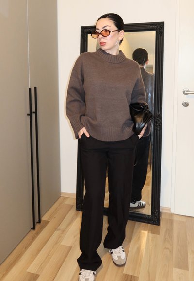 Mujer con suéter marrón, pantalones negros, zapatillas blancas y gafas de sol con lentes marrones, de pie con las manos en los bolsillos sosteniendo un bolso negro cerca de un espejo de cuerpo entero.