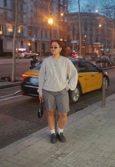 Sudadera gris, pantalones cortos grises, zapatos de plataforma negros, calcetines blancos. Bolso negro con asa naranja. Gafas de sol y joyería sencilla. Entorno urbano.