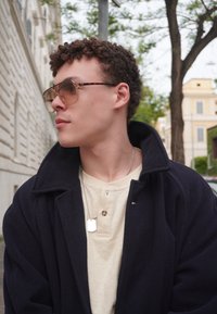 Casaco de lã preto, camisola henley bege claro, colar de chapa prateada, e óculos de sol grandes em padrão tartaruga; cabelo encaracolado, vista de perfil.