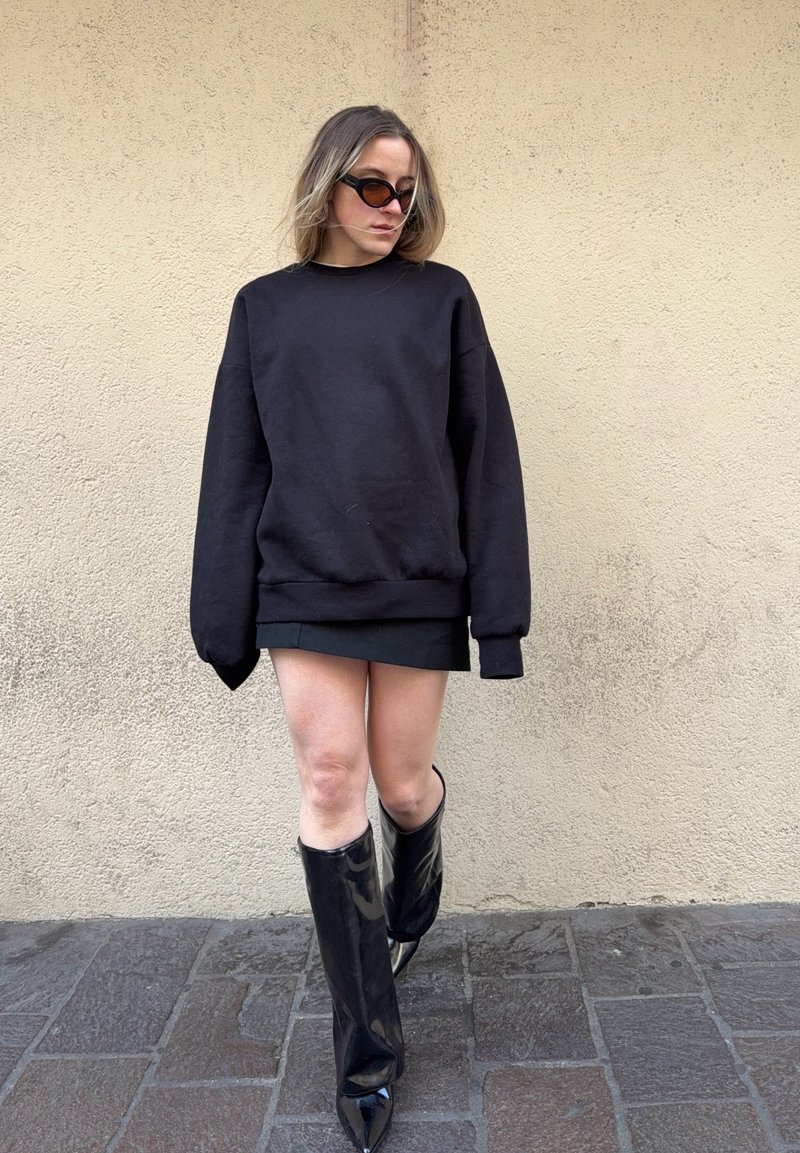 Kvinde iført oversized sort sweater, kort sort nederdel, knæhøje blanke sorte støvler og runde solbriller, stående op ad en struktureret beige væg.