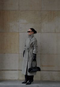 Šedý vlněný trenčkot s vysokým límcem, páskem v pase a přehnaně velkými knoflíky; doplněný černými koženými rukavicemi a kozačkami na vysokém jehlovém podpatku po kolena.