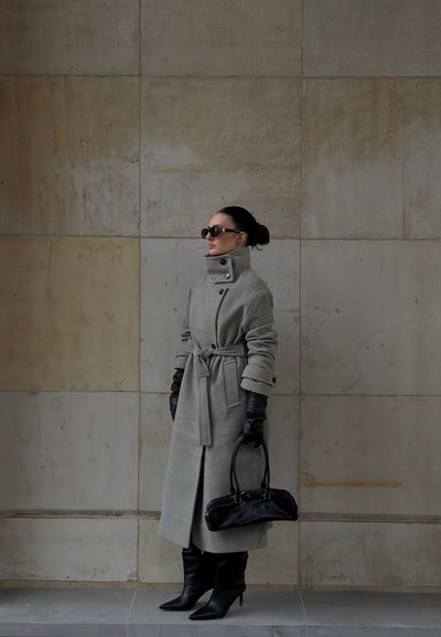 Abrigo tipo gabardina de lana gris con cuello alto, cintura ceñida con cinturón y botones grandes; combinado con guantes de cuero negro y botas de tacón fino hasta la rodilla.