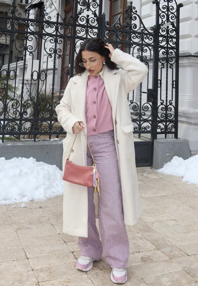 Mujer con abrigo largo blanco, cárdigan rosa, pantalones anchos lavanda y zapatillas, sosteniendo un bolso rosa, de pie junto a una puerta ornamental negra con nieve cerca.