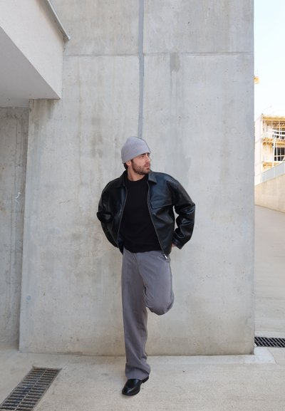 Hombre con gorro gris, chaqueta de cuero negra y pantalones grises, de pie sobre una pierna junto a una pared de hormigón en un entorno urbano.