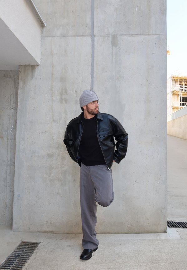 Hombre con gorro gris, chaqueta de cuero negra y pantalones grises, de pie sobre una pierna junto a una pared de hormigón en un entorno urbano.