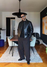 Sort skinn bomberjakke over en svart genser, kombinert med vide mørke bukser og blanke svarte sko. Moderne innendørs setting.