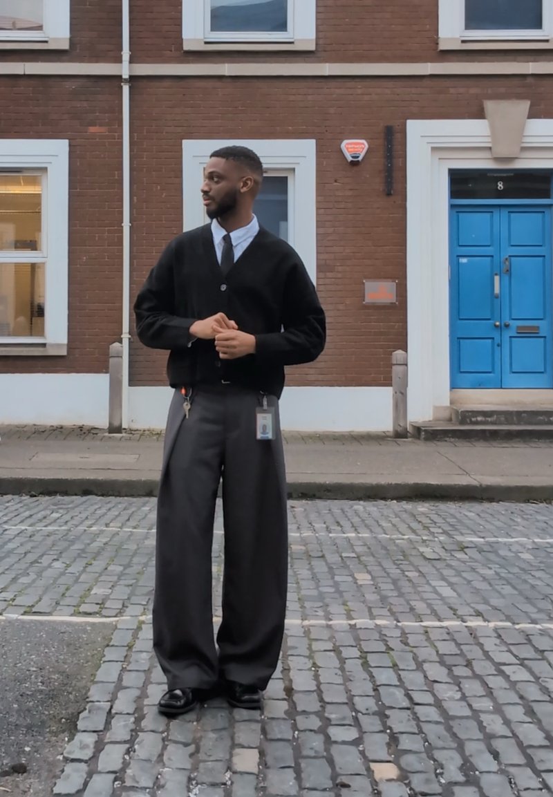 Mann trägt eine schwarze Strickjacke, ein weißes Hemd, eine Krawatte und lockere graue Hosen. Er hält Schlüssel und einen Ausweis in der Hand und steht auf Kopfsteinpflaster. Hinter ihm ist eine leuchtend blaue Tür.