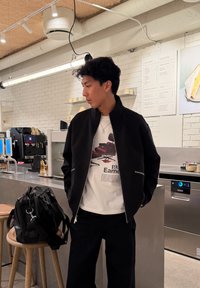 Jeune homme en veste noire et t-shirt graphique debout près d'un comptoir de café avec un sac noir sur un tabouret en bois à proximité.