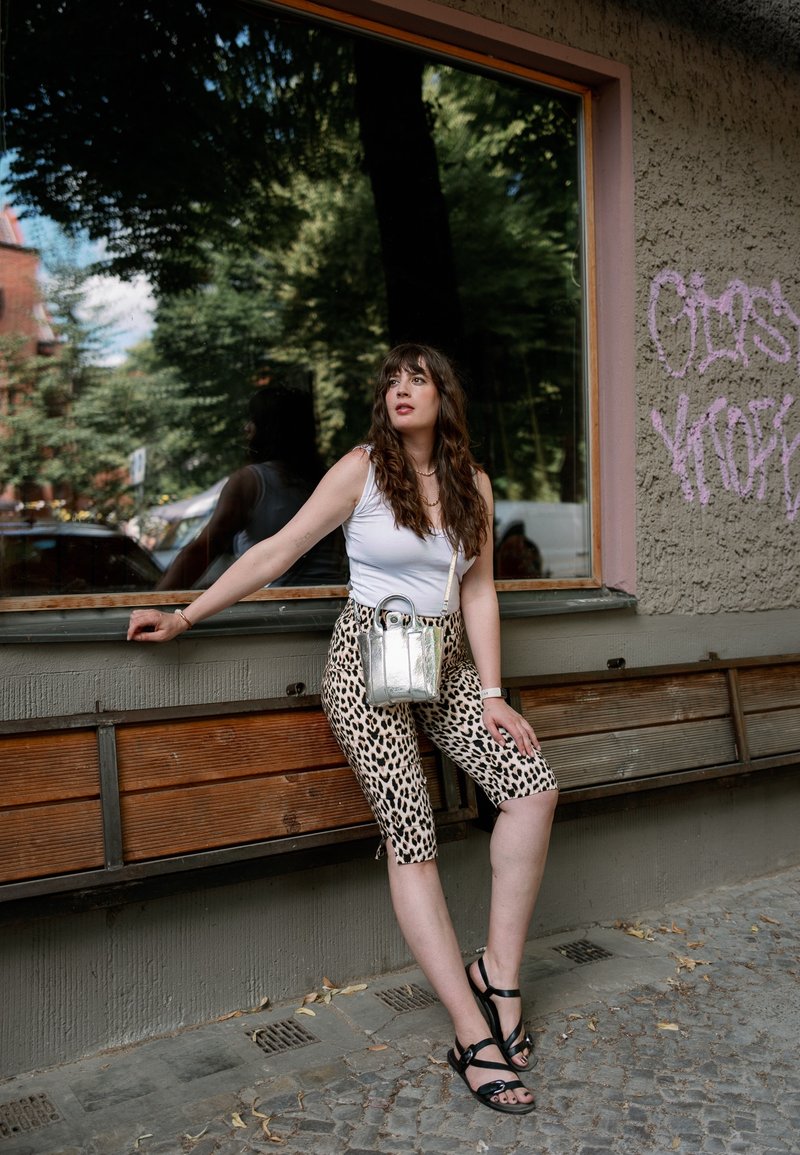 Femme en haut blanc et pantalon à motif léopard, assise sur un banc en bois devant un bâtiment avec une grande fenêtre réfléchissante.