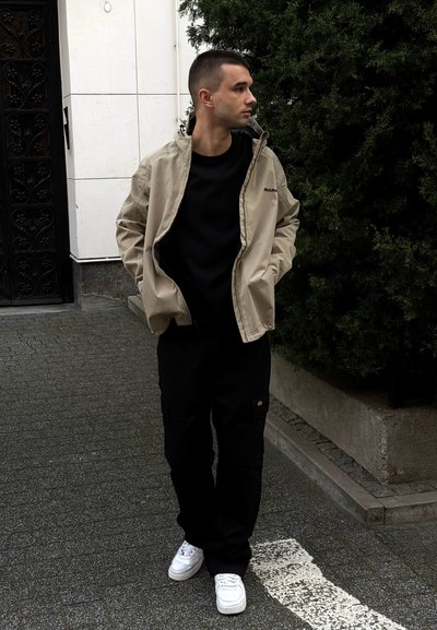 Chaqueta ligera beige sobre una camisa negra, combinada con pantalones cargo negros y zapatillas blancas; de pie sobre un pavimento de piedra cerca de vegetación.