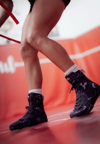 Črni in vijolični športni čevlji z visokimi zgornji deli, mrežasti in gumijasti materiali, teksturirana površina, nošeni z belimi gležnjarskimi nogavicami. Rdeča podlaga.