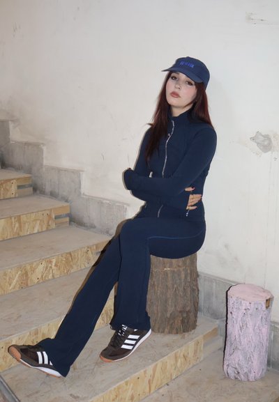 Mono atlético azul marino con cremallera, combinado con zapatillas marrones con rayas blancas. Modelo sentado en un taburete de madera contra un fondo sencillo.