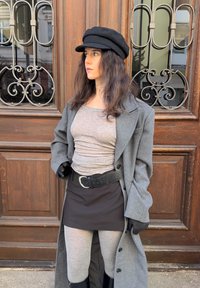 Manteau gris, ceinture noire, et pull associés à une jupe noire et des leggings gris ; portant une casquette noire et des gants, debout devant une porte en bois.