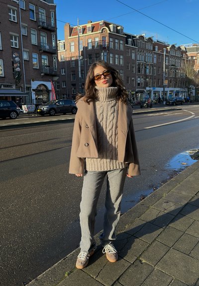 Abrigo beige de doble botonadura sobre un suéter de punto grueso con cuello vuelto, jeans gris de pierna ancha y zapatillas gris con suelas marrones. Gafas de sol grandes. Entorno urbano.