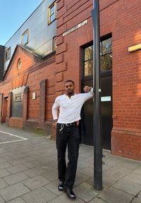 Homme en chemise blanche à manches longues et pantalon noir appuyé contre un poteau noir à l'extérieur d'un bâtiment en briques rouges par une journée ensoleillée.