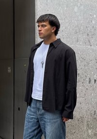 Černá košile na knoflíky přes bílé tričko, doplněná světle modrými džínami. Košile má volný střih a hladkou texturu, s minimálním designem.