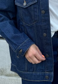 Τζάκετ από τζιν σε σκούρο μπλε με χρυσές ραφές, με ασημένια κουμπιά και μπροστινή τσέπη. Χέρι να κρατά την περιοχή με τα κουμπιά.