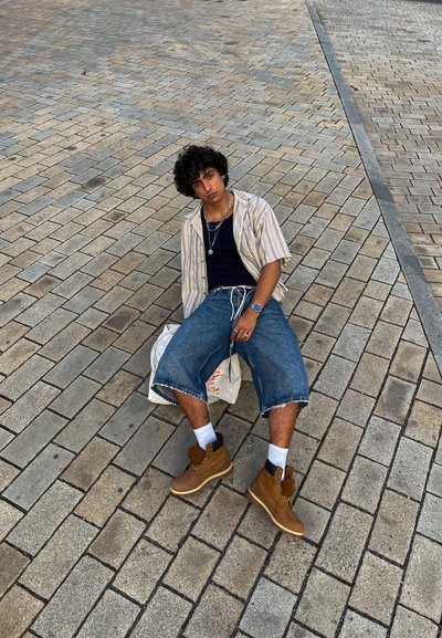 Una persona está sentada en un pavimento de piedra con una camisa de rayas de mangas cortas, una camiseta oscura, pantalones cortos de mezclilla y botas marrones con calcetines blancos, sosteniendo una bolsa.