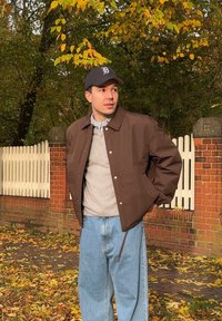 Geacă maro cu guler, cămașă gri dedesubt și blugi albastri din denim largi. Înconjurat de frunze de toamnă și un gard alb.